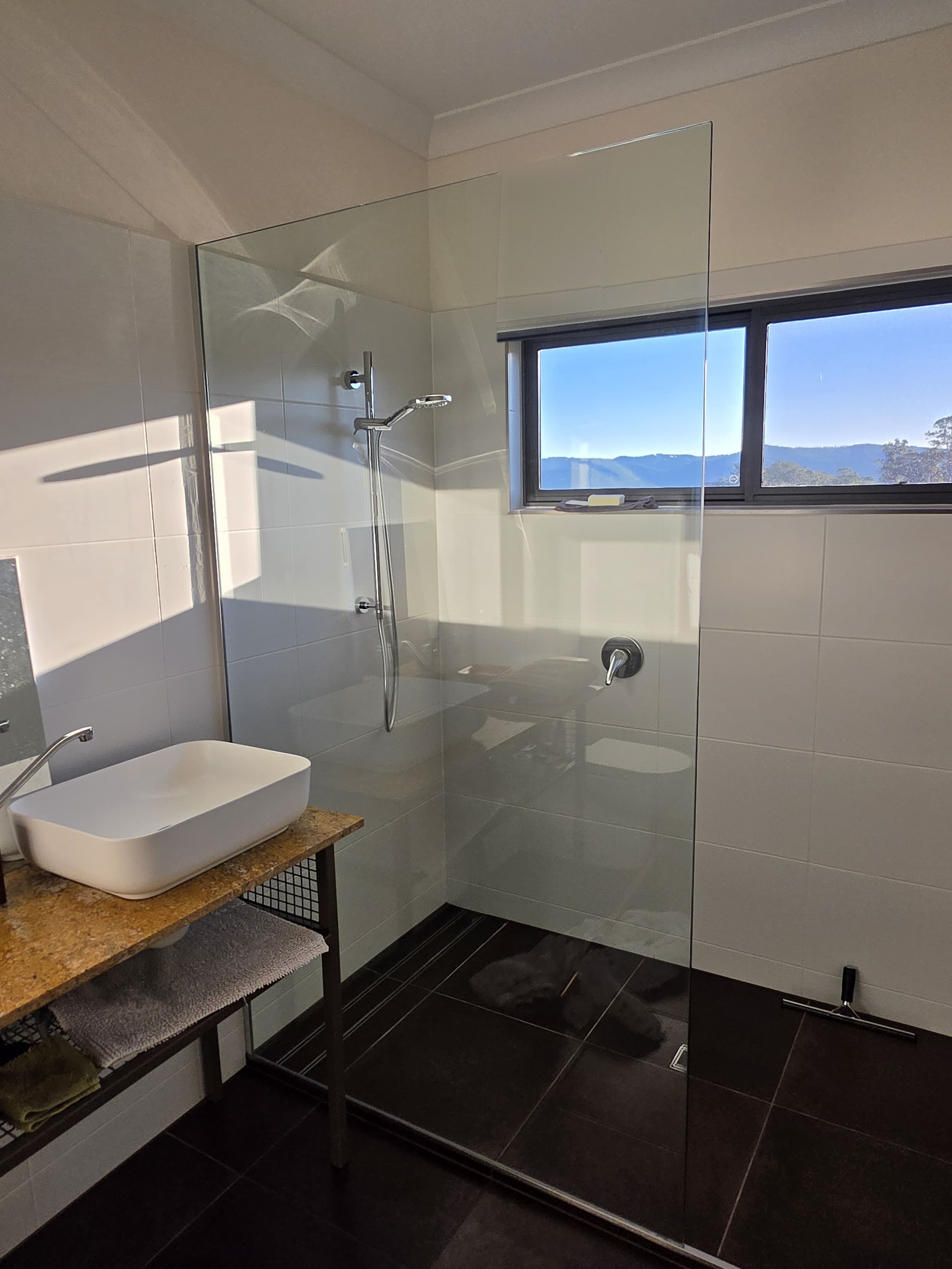 Bathroom with a glass shower screen, a white vessel basin on a timber top, dark floor tiles, and a window looking out to hills.