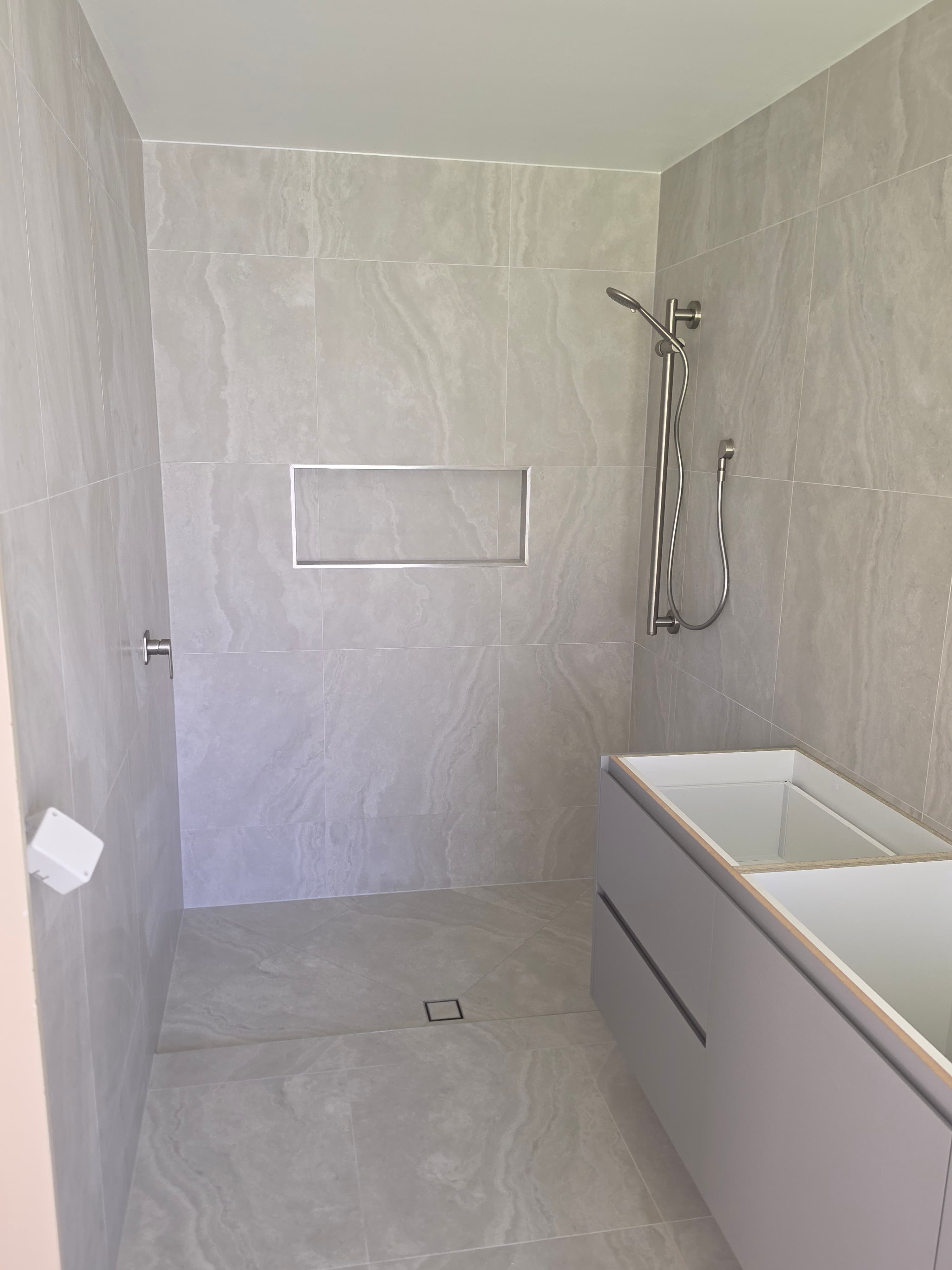 Walk-in shower lined with large grey tiles, with a recessed niche, a floor waste, and a grey vanity with a white bath on the right.