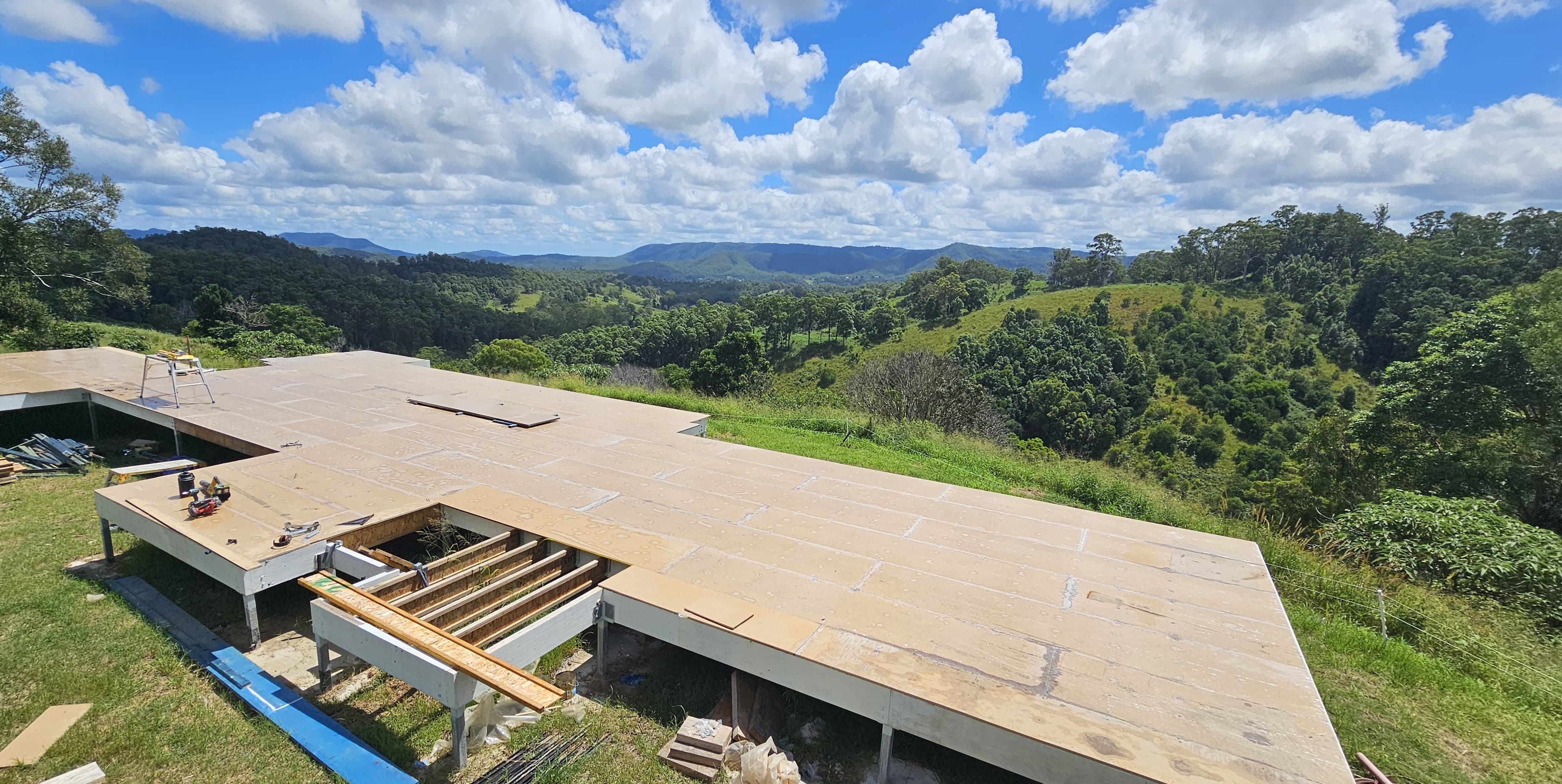Plywood sheets on a steel and timber floor platform under construction, with framing open in places, on a grassy hillside with bush and mountains behind.