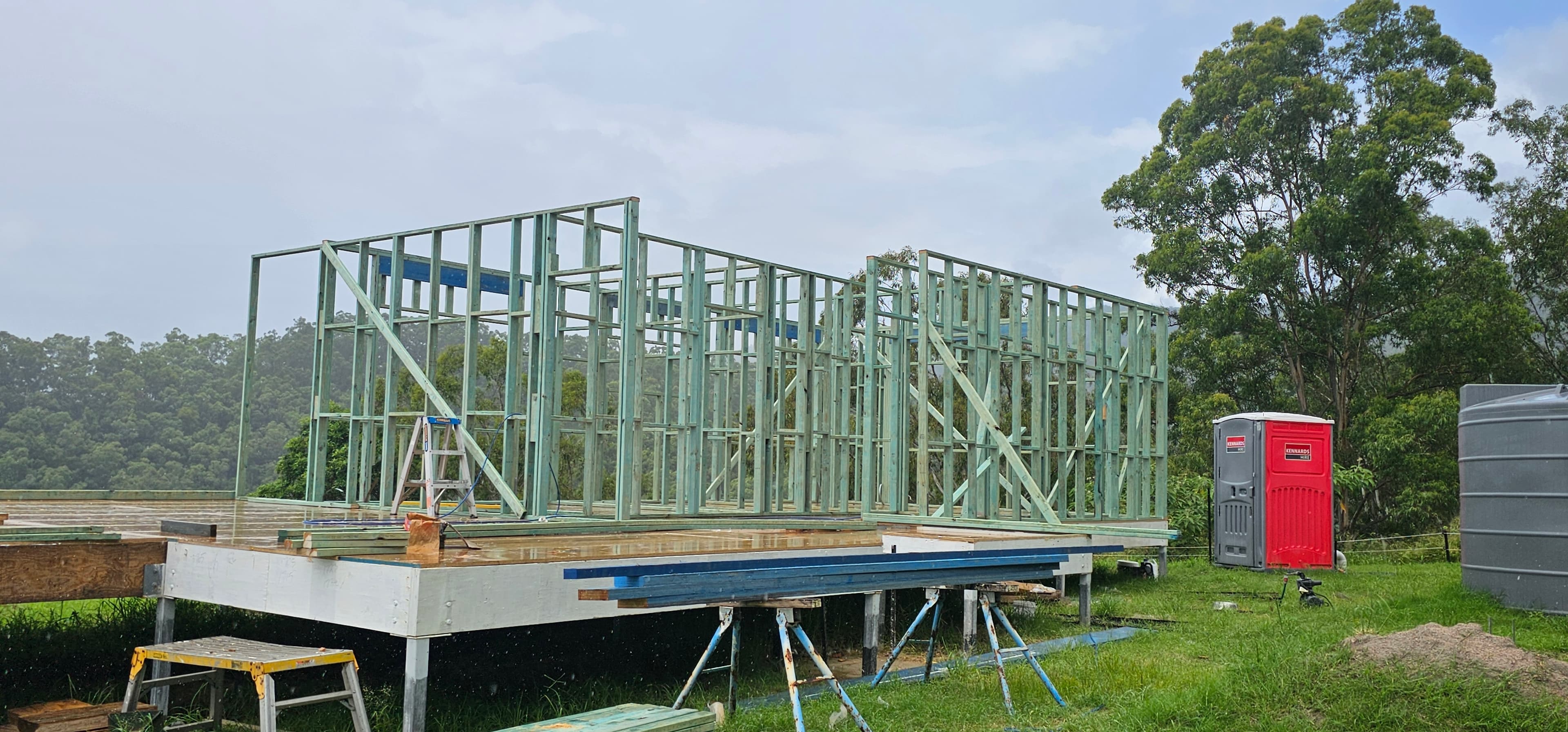 Timber wall frames standing on a raised floor platform, with a ladder, a red portable toilet, and a water tank to the right, and trees behind.