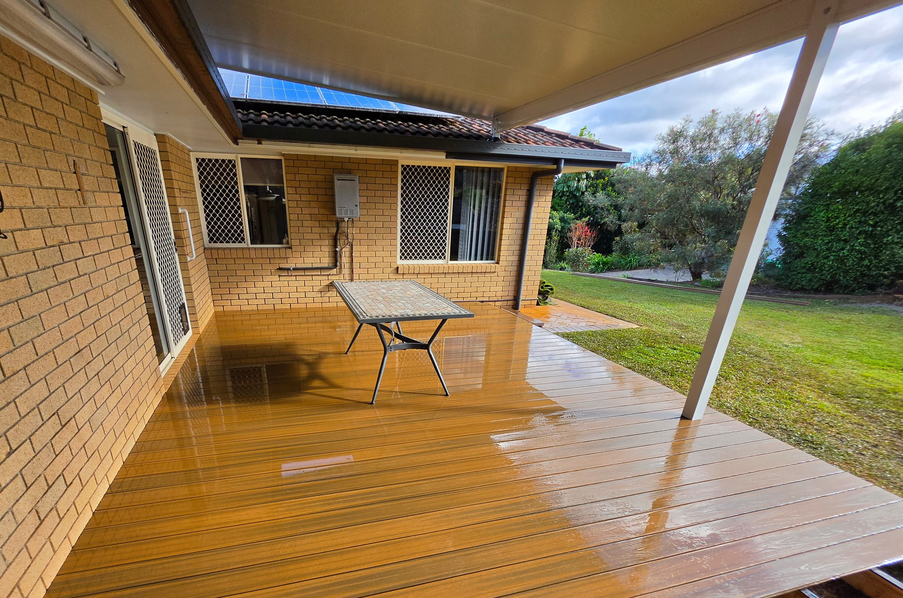 Covered deck on a brick house, with boards under a roof, an outdoor table, and lawn to the side.