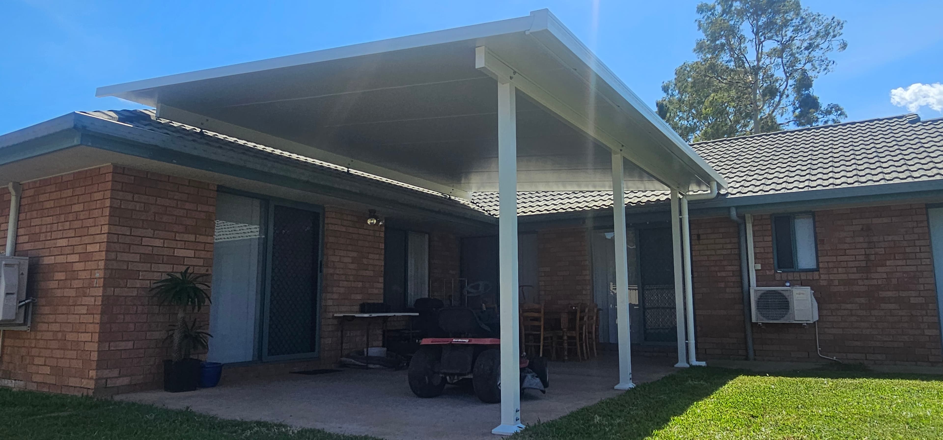White metal patio roof on white posts attached to a brick house, with a concrete slab underneath, lawn in front, and a ride-on mower and outdoor furniture under the roof.