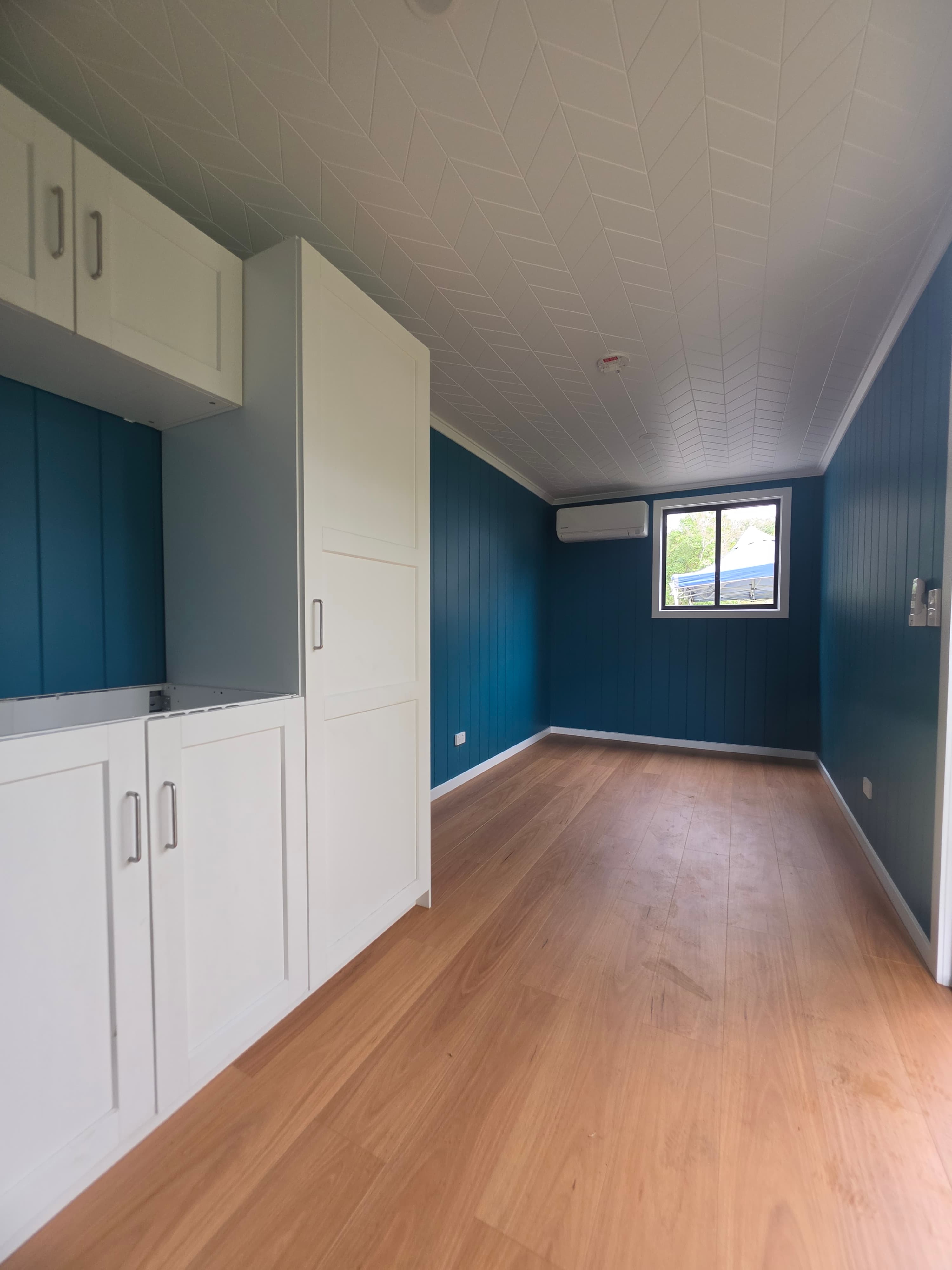 Kitchen end of the same interior, with white cabinets, one opening without a door, blue walls, a window, a wall-mounted air conditioner, timber floor, and a white geometric ceiling.