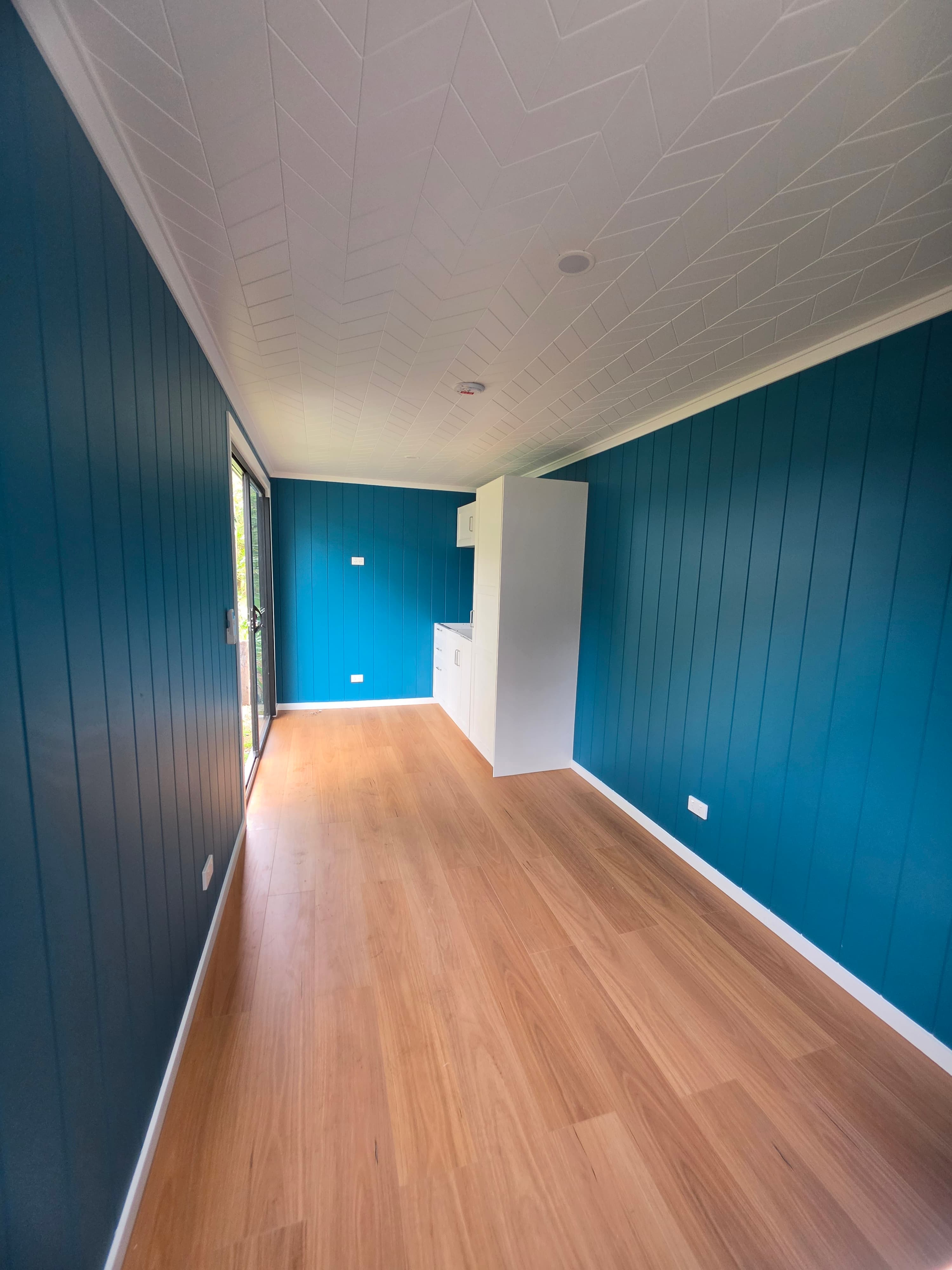 Long narrow finished interior with blue vertical wall panels, a white geometric ceiling, timber floor, white kitchen cabinetry at the far end, and a sliding door on the left.