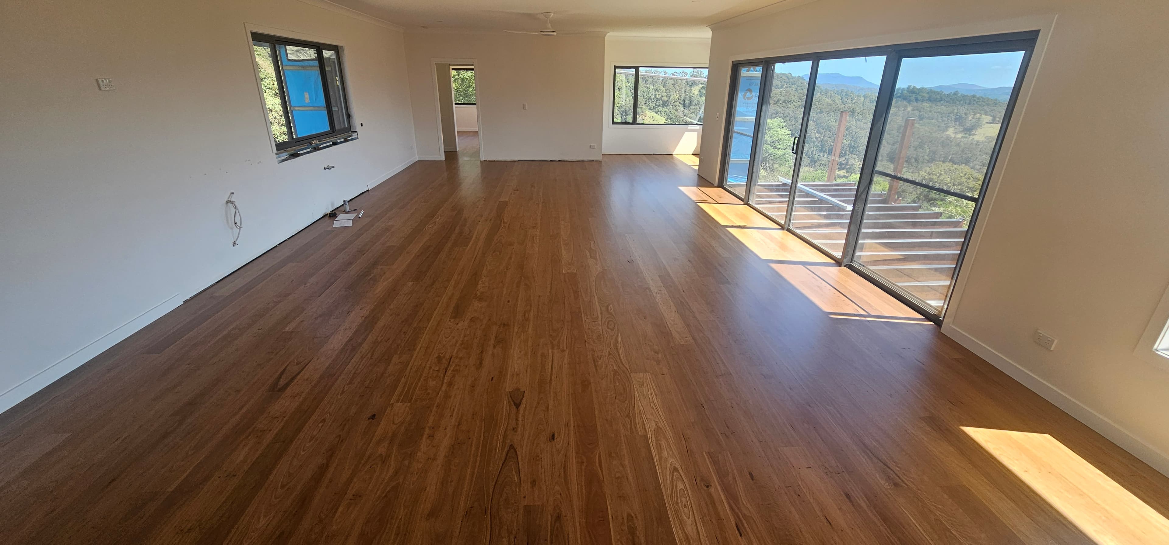 Finished living area with timber floorboards, white walls, a ceiling fan, windows, and sliding glass doors opening onto a deck with hills beyond.