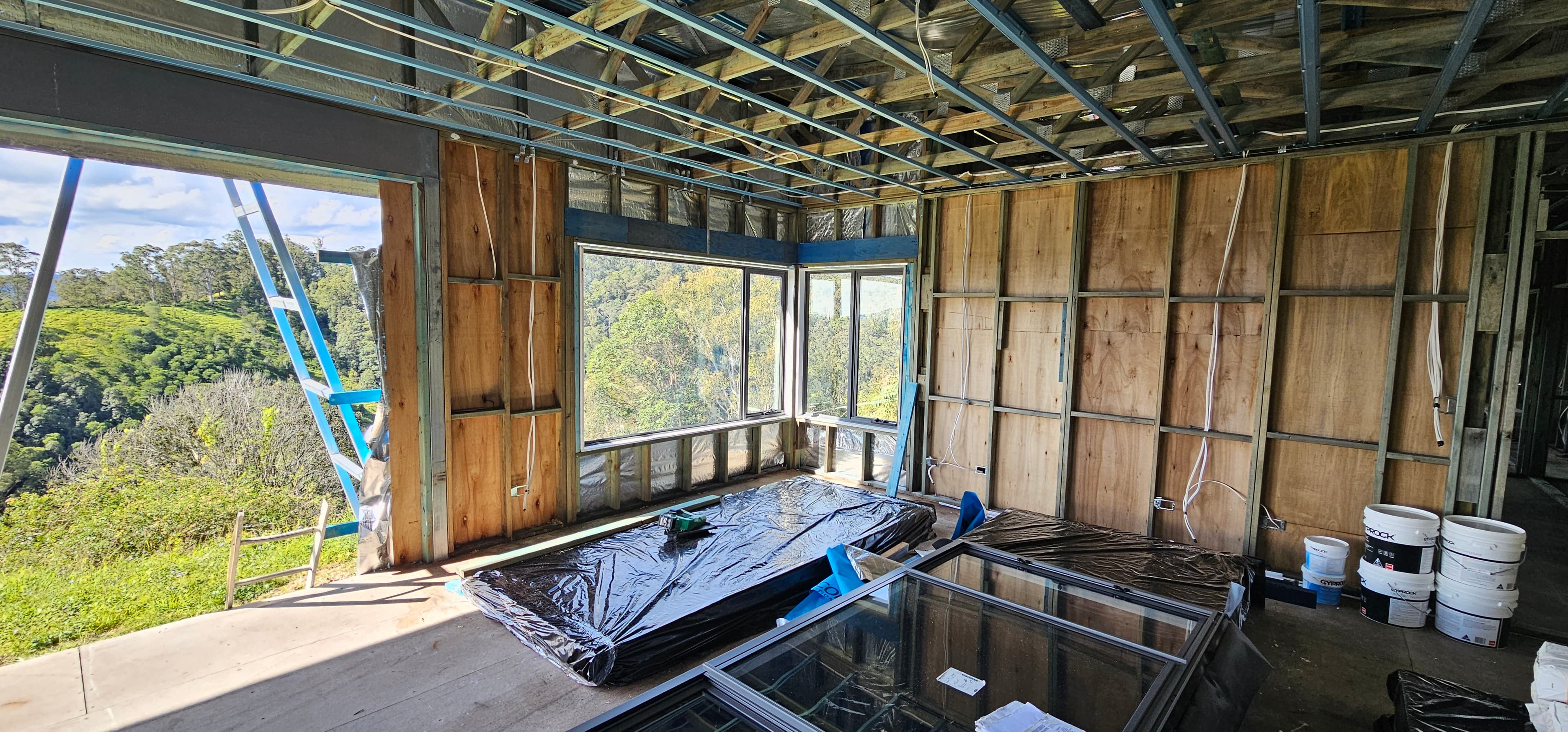 Room under construction with timber and steel wall frames, plywood sheets, windows looking onto bush and hills, exposed ceiling battens, stacked glass door frames in the foreground, a ladder, and paint buckets.